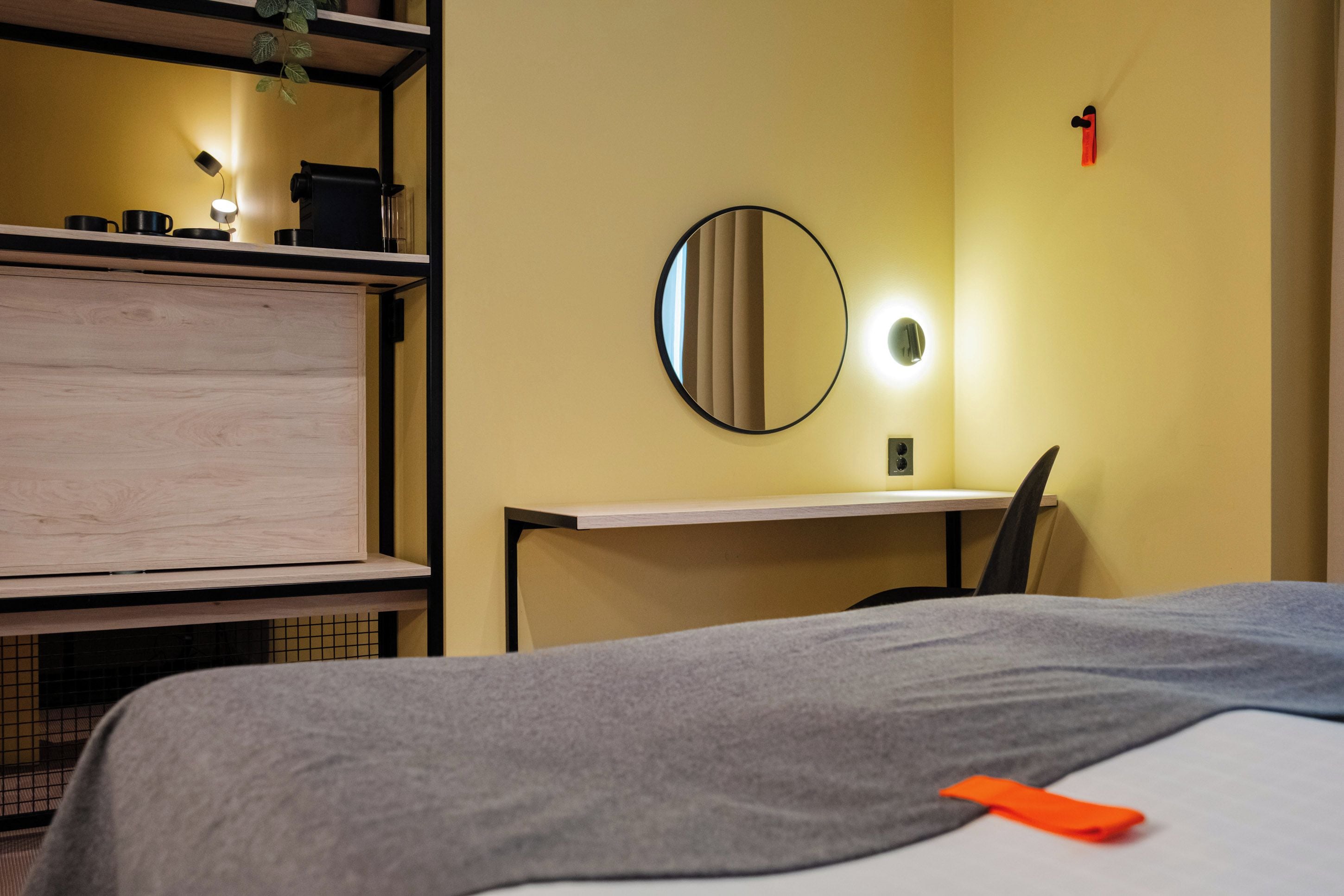 Et hotelværelse med en seng, et skrivebord, et spejl og en reol med ting.