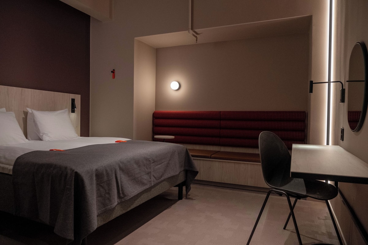 A hotel room with a bed, bedside table, chair, and a red bench against the wall.