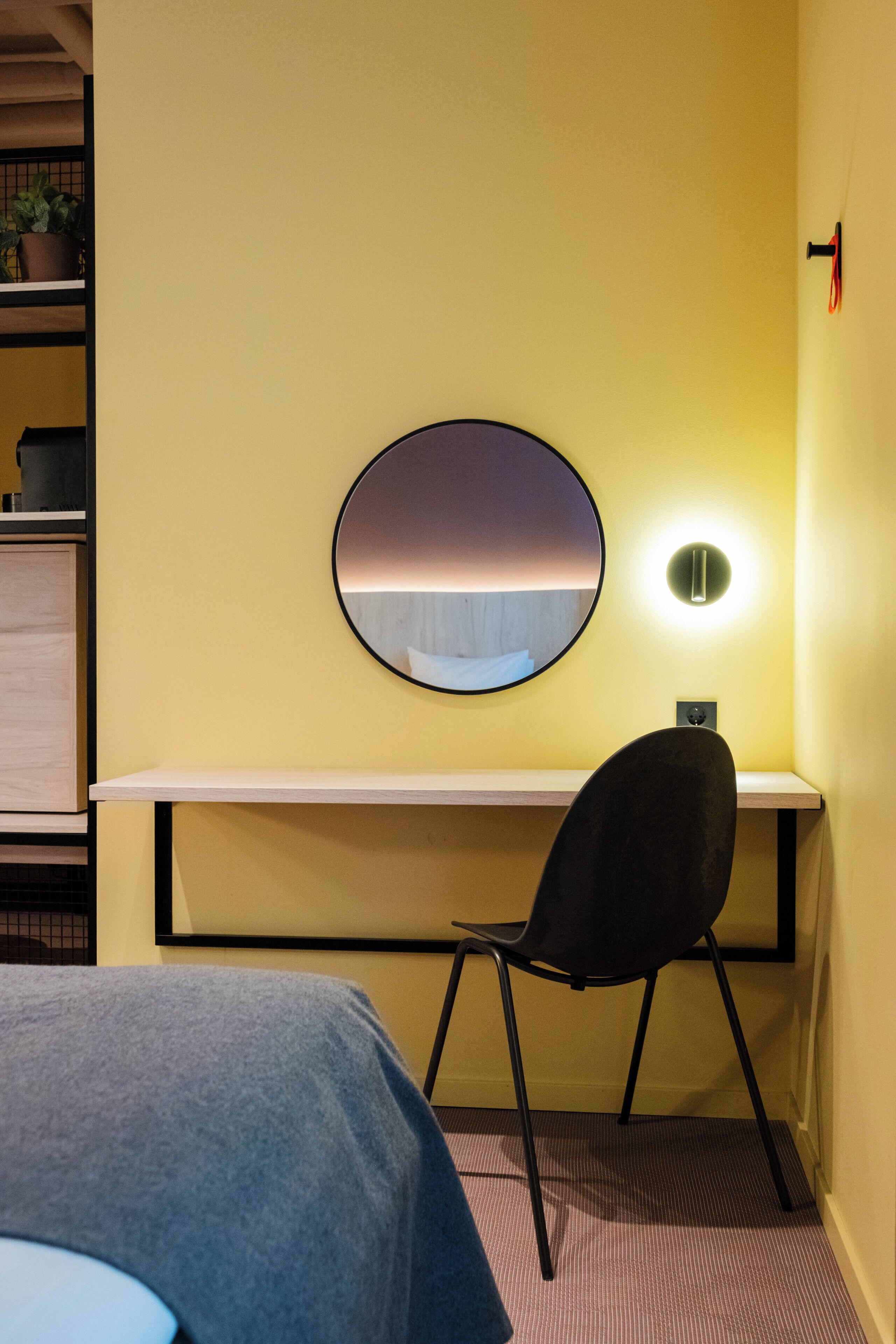 Een kamer met een bed, een bureau met stoel, een ronde spiegel en een wandlamp.