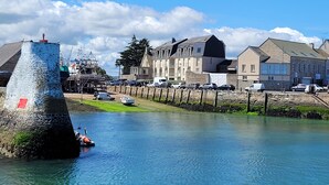 A waterfront area with a stone tower, a row of parked cars, and buildings with varying architectural styles. - 2-room garage facing with panoramic view of the sea and TATIHOU island (UNESCO) (Saint-Vaast-la-Hougue)
