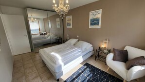 A bedroom with a bed, a white armchair, a bedside table with a lamp, a chandelier, and a poster on the wall. - 2-room garage facing with panoramic view of the sea and TATIHOU island (UNESCO) (Saint-Vaast-la-Hougue)