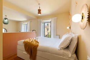 A bedroom with a bed, a bedside table, a window with curtains, and a wall-mounted light fixture.