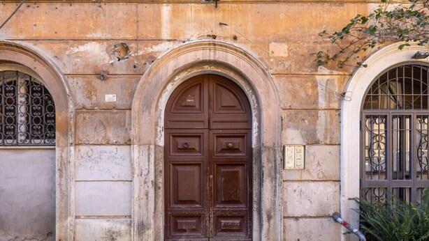 Un bâtiment historique doté d’une grande porte en bois richement décorée et de deux fenêtres en arc surmontées de grilles métalliques.