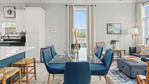 Eine moderne Küche mit weißen Schränken, ein Essbereich mit blauen Stühlen und einem Glastisch sowie ein Wohnzimmer mit einem grauen Sofa und Blick ins Freie.
