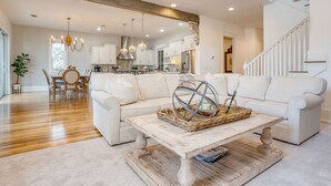 A spacious living room with a white sofa, wooden coffee table, and a dining area with a wooden table and chairs. - Sleeps 18 | Luxe 30A Beach Home w/ Resort Pool + Outdoor Fireplace (Santa Rosa Beach)