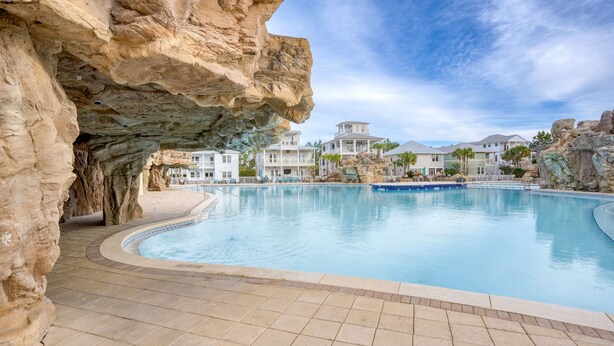 A swimming pool with a rocky cave-like structure on one side, surrounded by modern buildings with a clear sky above. - Sleeps 18 | Luxe 30A Beach Home w/ Resort Pool + Outdoor Fireplace (Santa Rosa Beach)