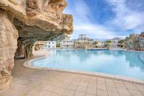 A swimming pool with a rocky cave-like structure on one side, surrounded by modern buildings with a clear sky above. - Sleeps 18 | Luxe 30A Beach Home w/ Resort Pool + Outdoor Fireplace (Santa Rosa Beach)