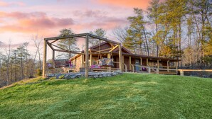 A wooden cabin with a porch and a stone foundation, surrounded by trees and a grassy hill. - Designer Retreat • Pool + Theater + Hot Tub • Near Pigeon Forge | Pink Pines (Sevierville)