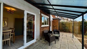 A covered patio with a table and chairs, a glass door leading to an interior room, and a brick wall. - Sandringham House Contractors, Families, Holidays (Cleethorpes)