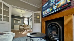 A living room with a fireplace, a television mounted above it displaying a Netflix interface, a round table with a remote, and a sofa.