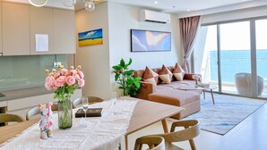 A modern living room with a dining area, featuring a sofa set, a dining table with a vase of flowers, and a large window with a view of the sea. - The Song Sunny Canopy (Ho Chi Minh City)