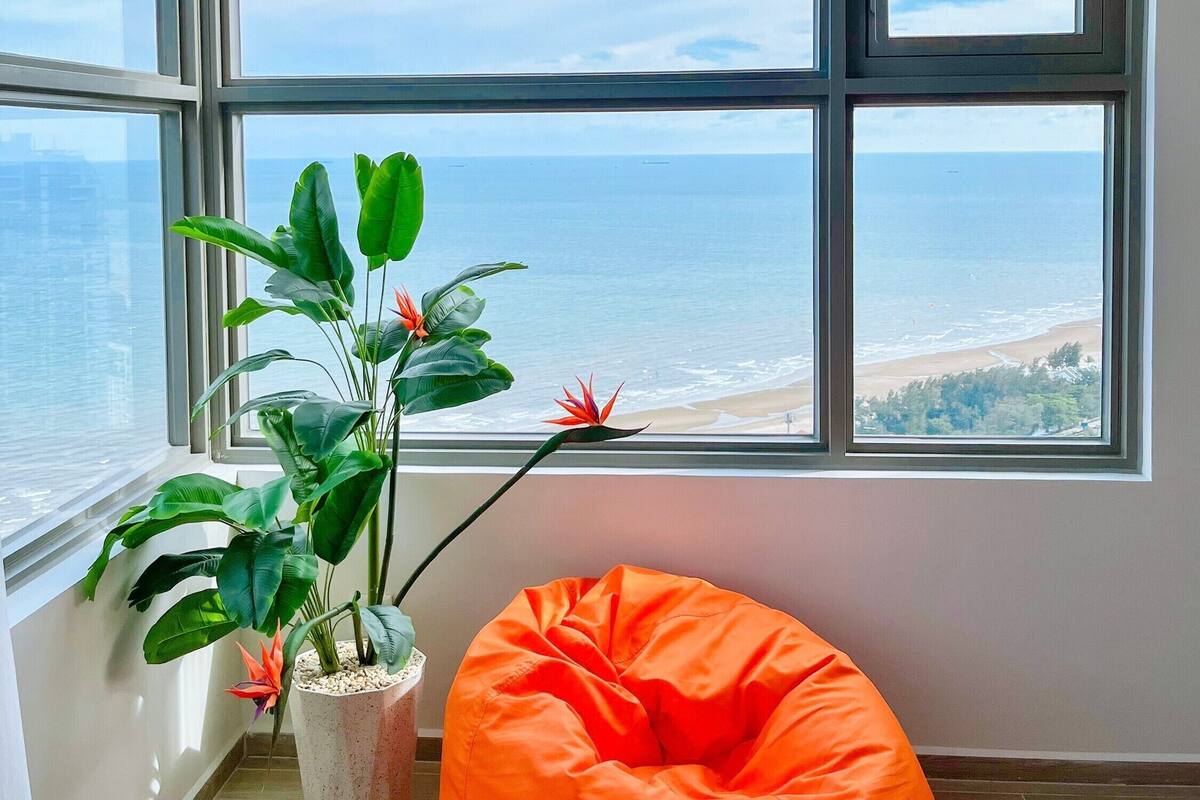 Una habitación con un ventanal grande con vistas a la playa, una planta en maceta y un sillón de mezcla naranja.