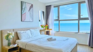 A neatly arranged bedroom with a large bed, a bedside table with a vase of flowers, a framed picture on the wall, and a view of the sea through a large window. - The Song Sunny Canopy (Ho Chi Minh City)