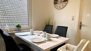 A dining area with a table set for two, chairs, a clock on the wall, and a window with a view of the roof.