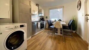 A compact kitchen with a washing machine, a dining area with a table and chairs, and a window with blinds.