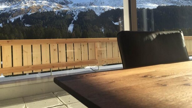 Een balkon met een houten hekwerk, een houten tafel en uitzicht op besneeuwde bergen.
