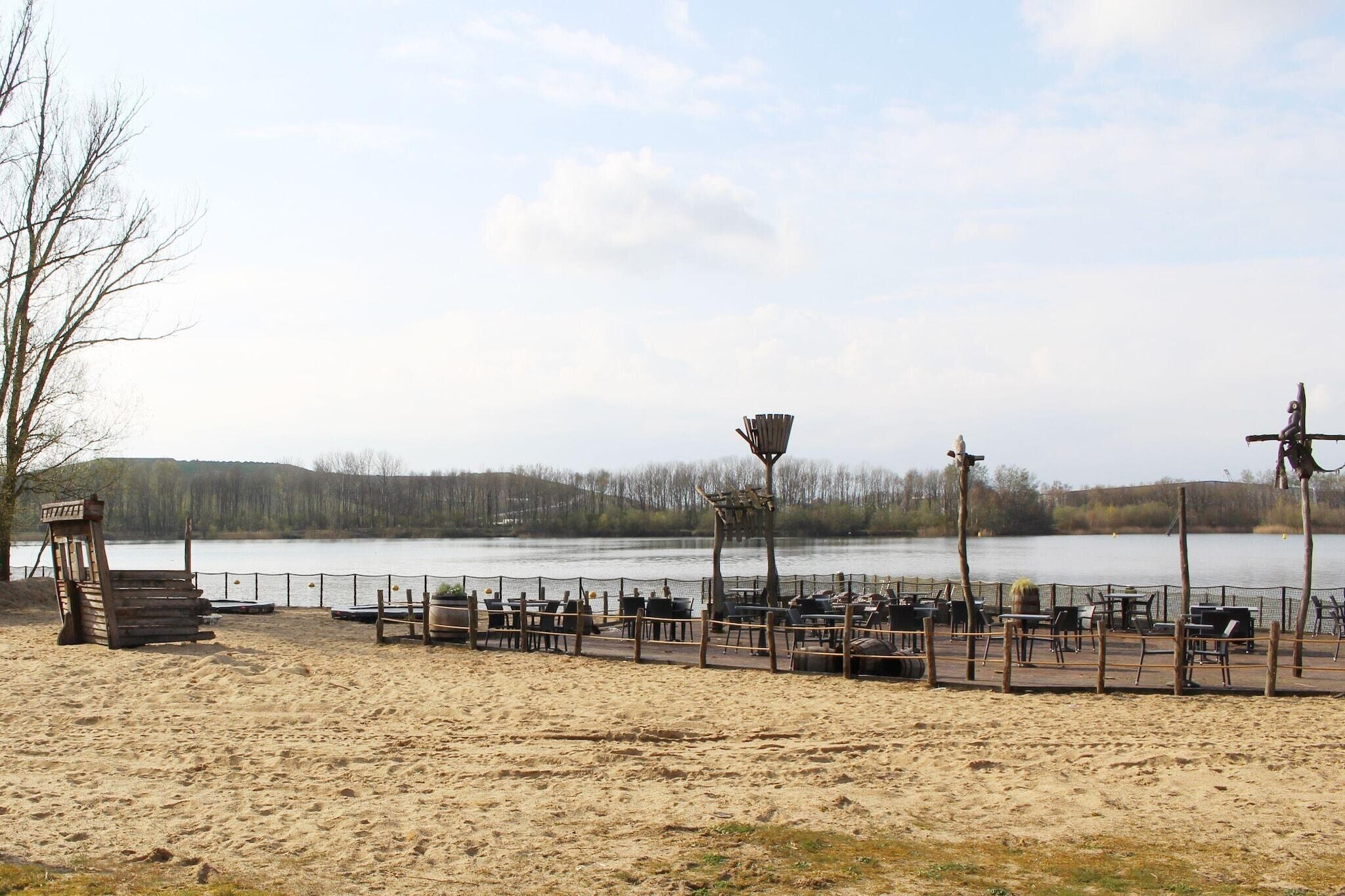 A lakeside area with a wooden deck, tables, and chairs, surrounded by a fence.