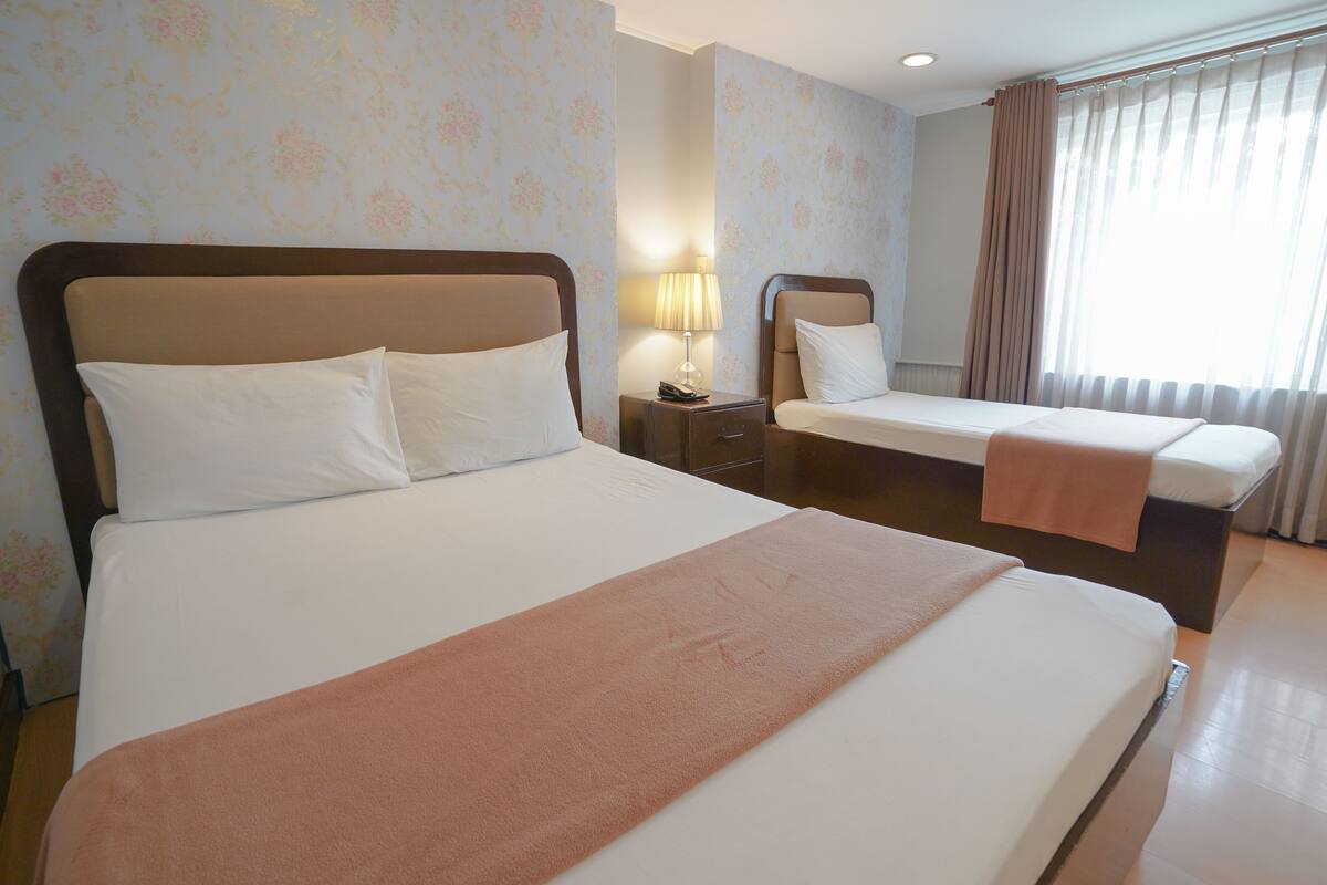 A hotel room with two beds, a nightstand, a lamp, and a window with curtains.