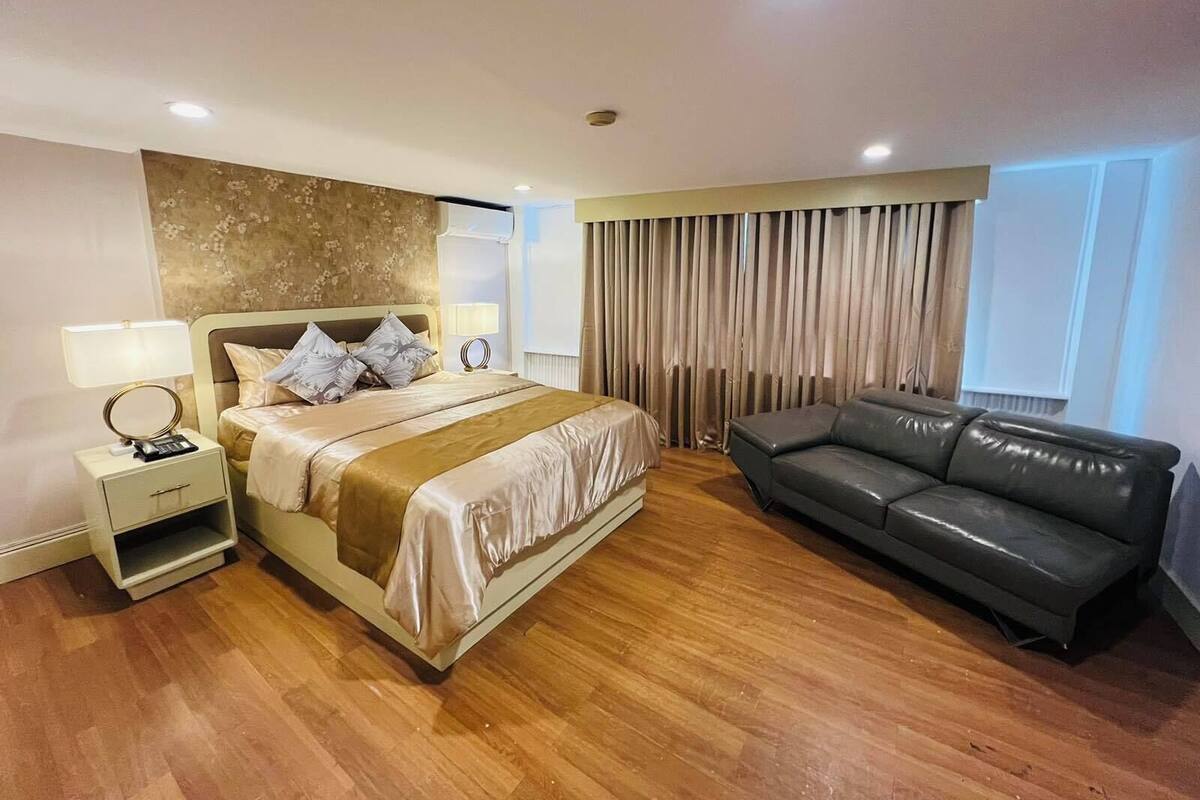 A hotel room with a bed, a nightstand, a lamp, a sofa, and a large window with curtains.