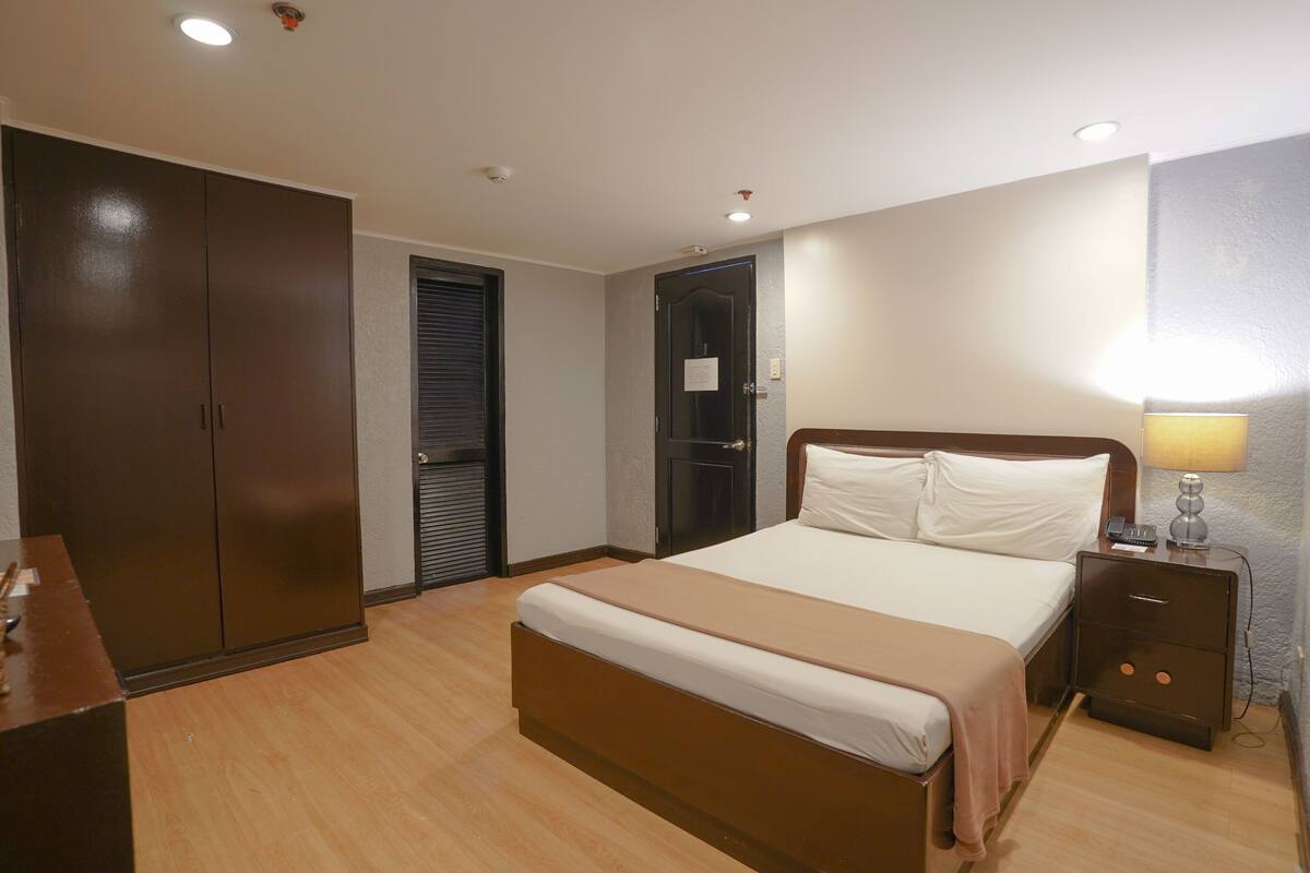 A bedroom with a large bed, a wardrobe, a bedside table with a lamp, and a door with a sign.