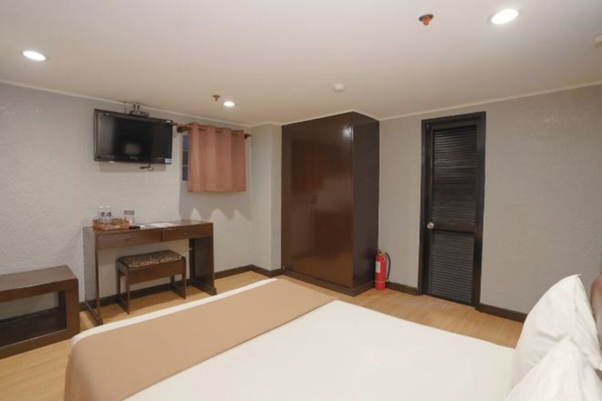 A hotel room with a bed, a desk with a chair, a television mounted on the wall, a wardrobe, and a fire extinguisher.