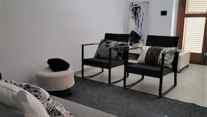 A modern living room with a sofa, two armchairs, a coffee table, and a small round ottoman. There is a painting on the wall and a window with blinds.