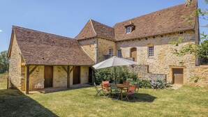 A stone building with a tiled roof, a covered outdoor seating area with a table and chairs, and a small garden.