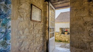 A stone-walled corridor with a wooden door leading to an outdoor area with a tiled roof and a potted plant.