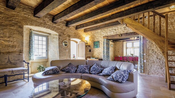 A rustic interior with a curved leather sofa, a glass coffee table, stone walls, exposed wooden beams, and a staircase.