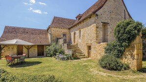 A stone house with a tiled roof, a covered patio with chairs, and a garden with flowers and shrubs.