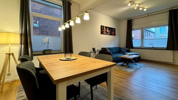 Un salon moderne avec une table à manger en bois, un canapé et une table basse. De grandes fenêtres sont présentes, ainsi que des rideaux, une lampe et un tableau accroché au mur.