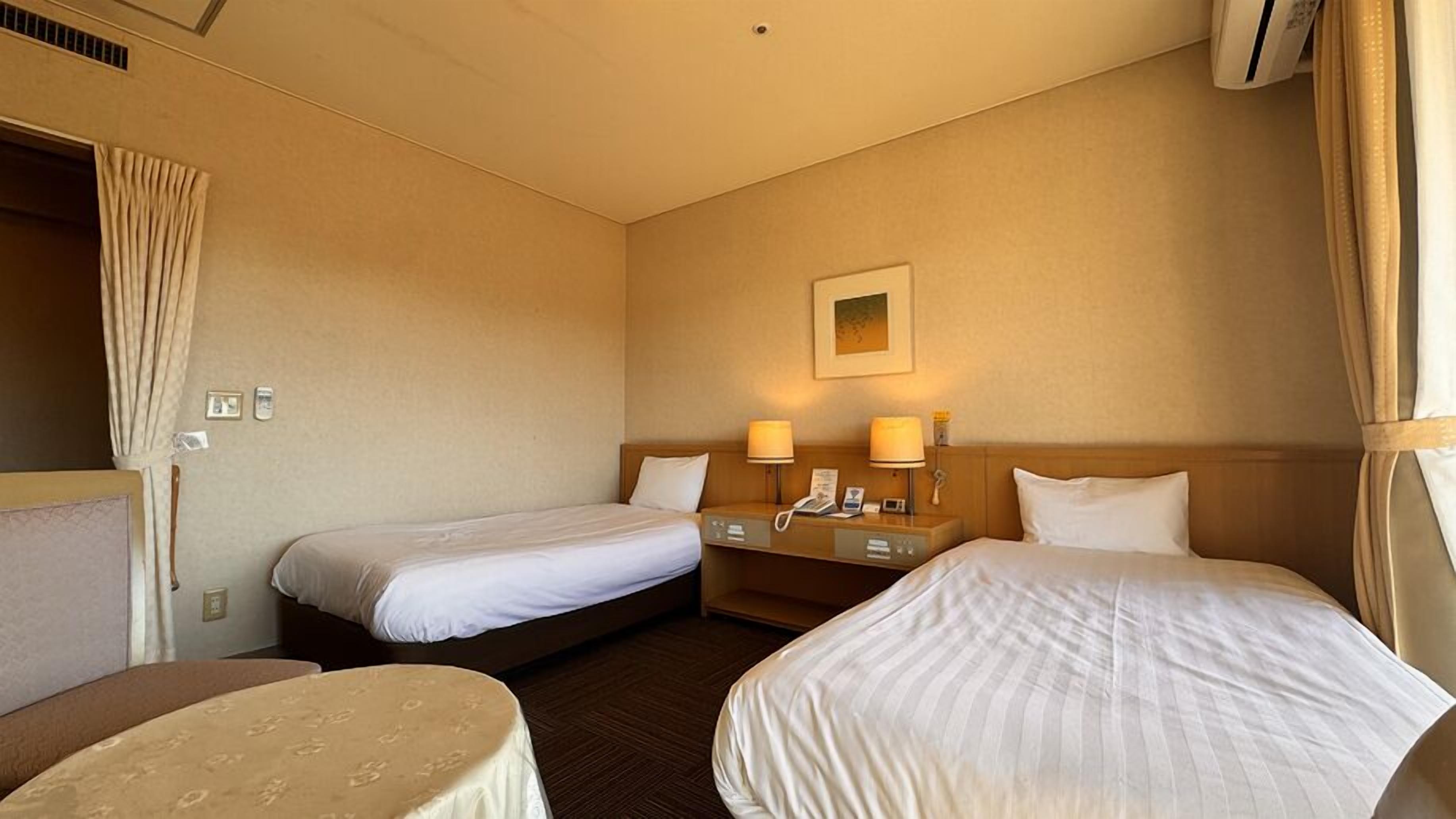 A hotel room with two single beds, a small table, a bedside table with lamps, and a wall-mounted picture.