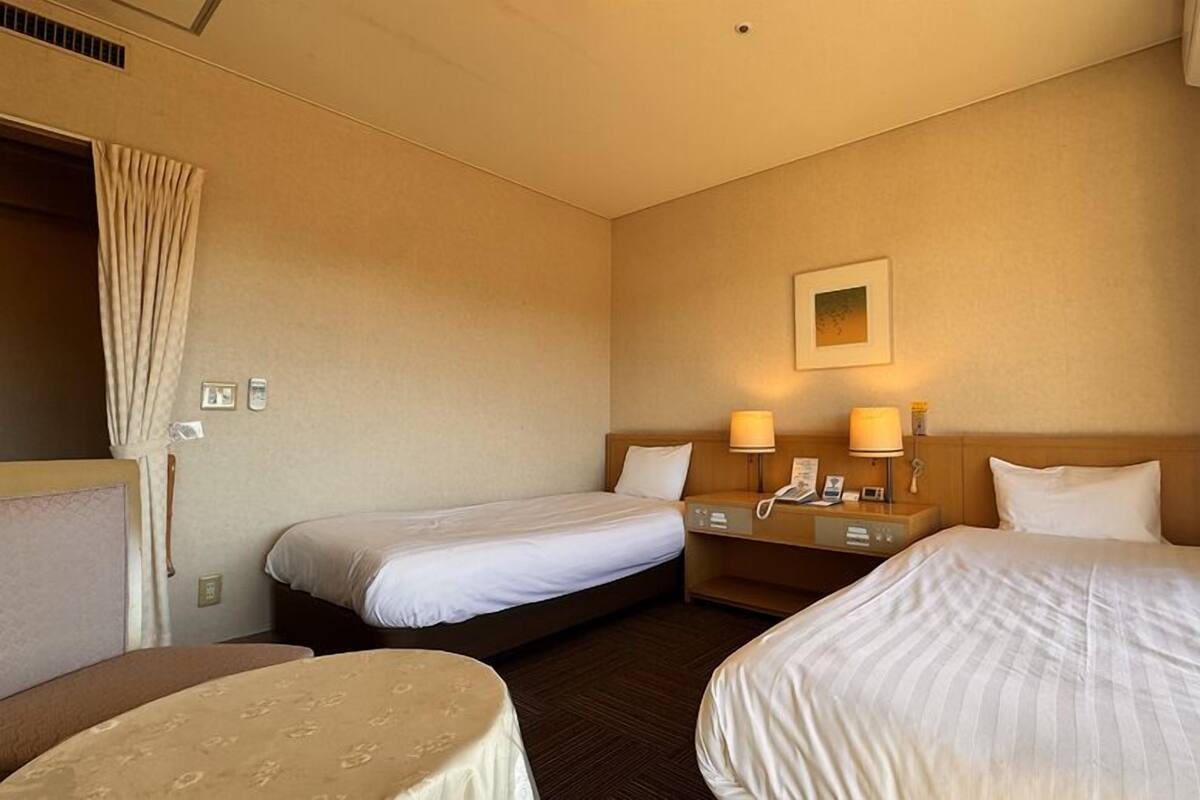 A hotel room with two single beds, a small table, a bedside table with lamps, and a wall-mounted picture.
