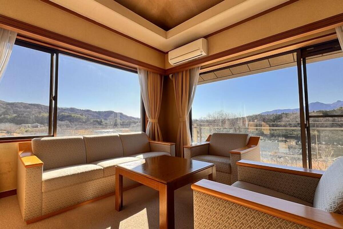 A room with a sofa, chairs, a table, large windows, and an air conditioning unit.