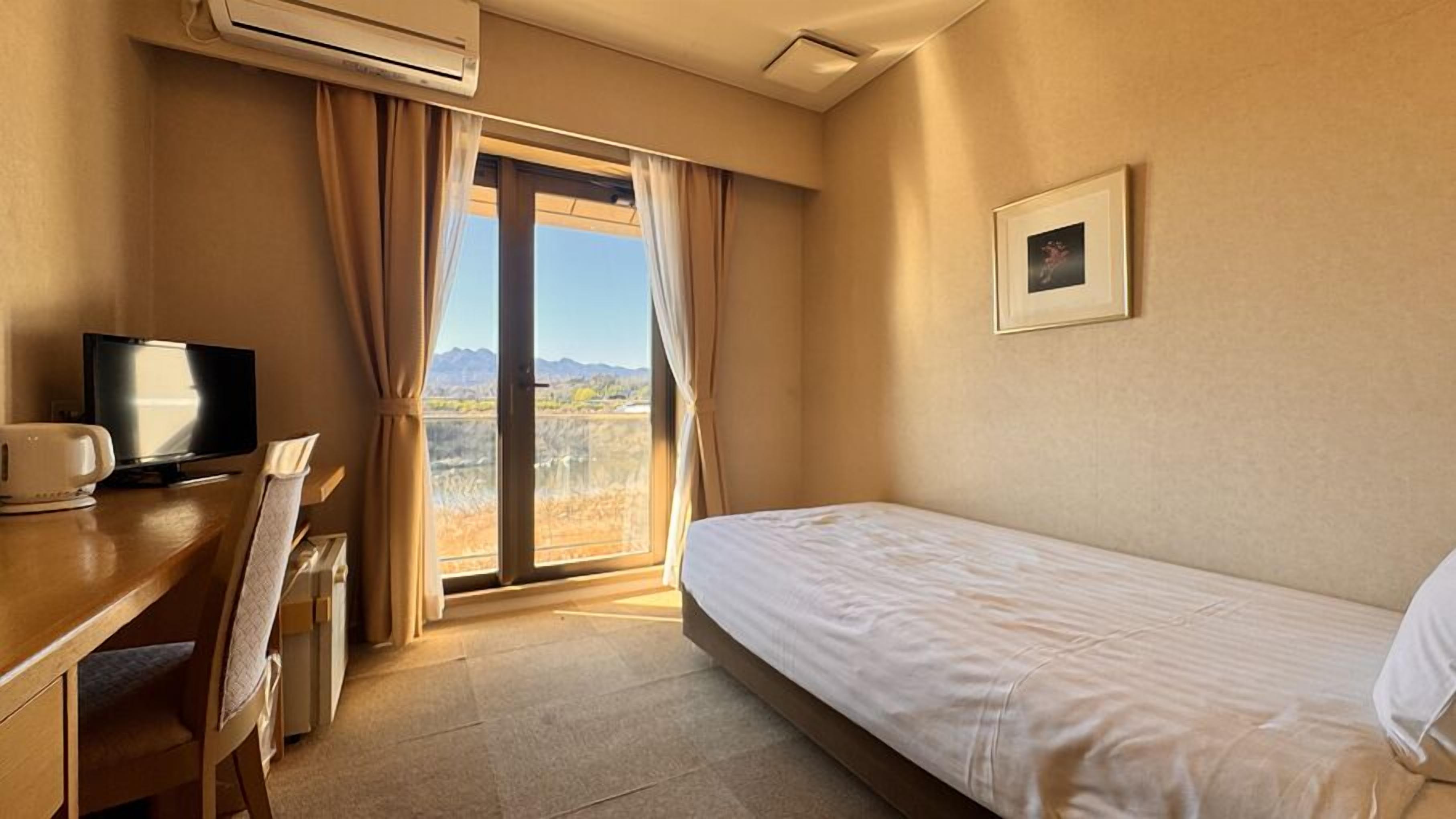 A hotel room with a bed, a desk with a television, an air conditioner, and a window with curtains.