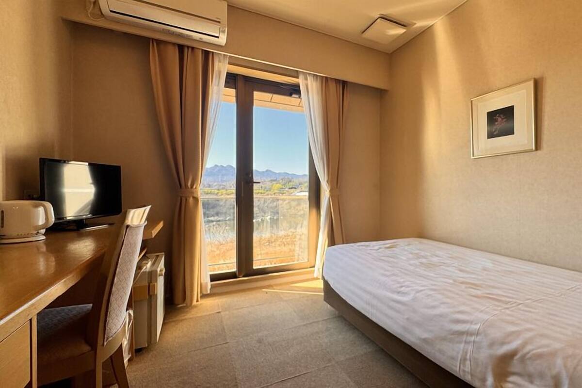 A hotel room with a bed, a desk with a television, an air conditioner, and a window with curtains.