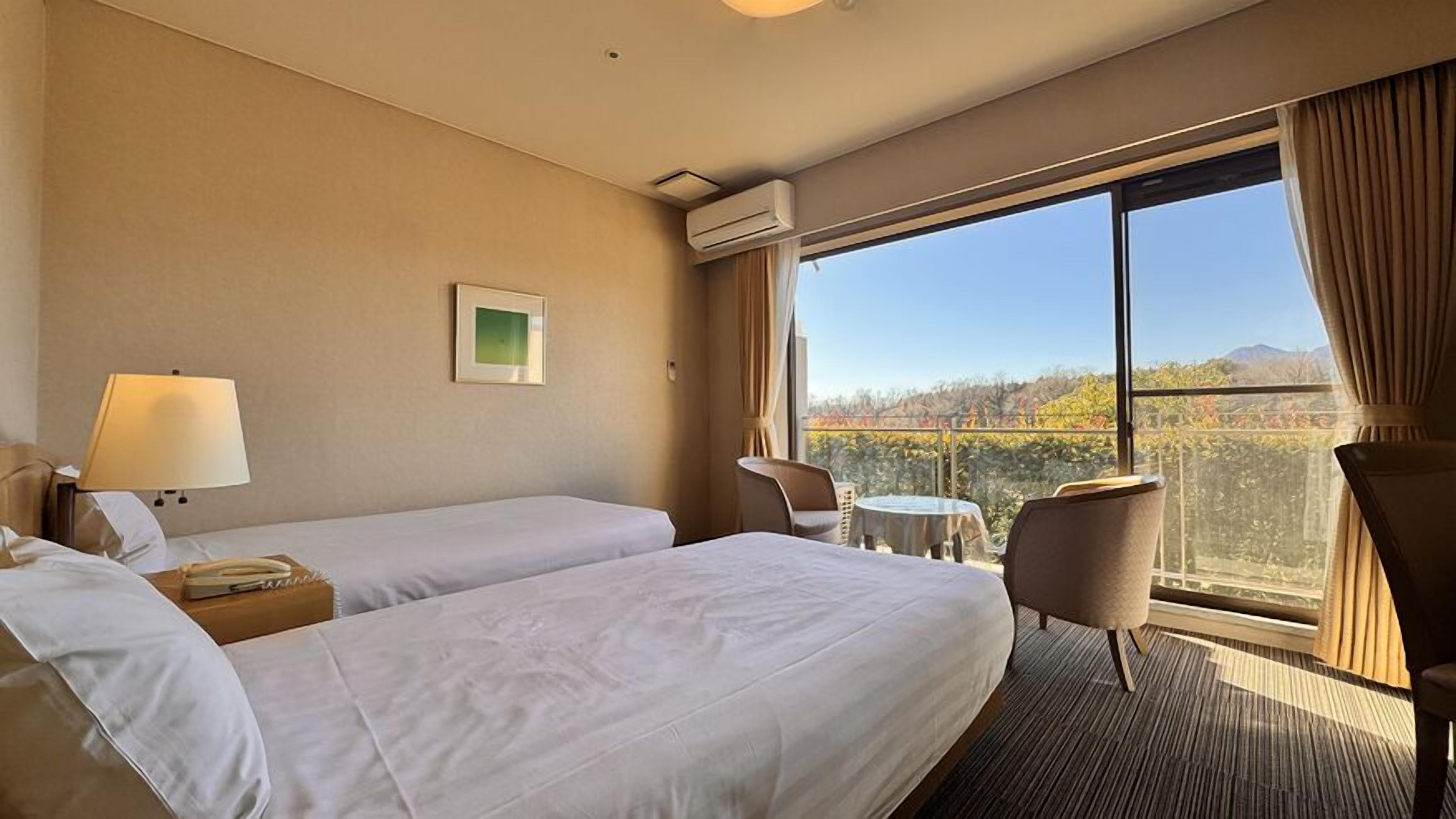 A hotel room with two beds, a desk, a chair, a small table, and a large window offering a view of trees and mountains.