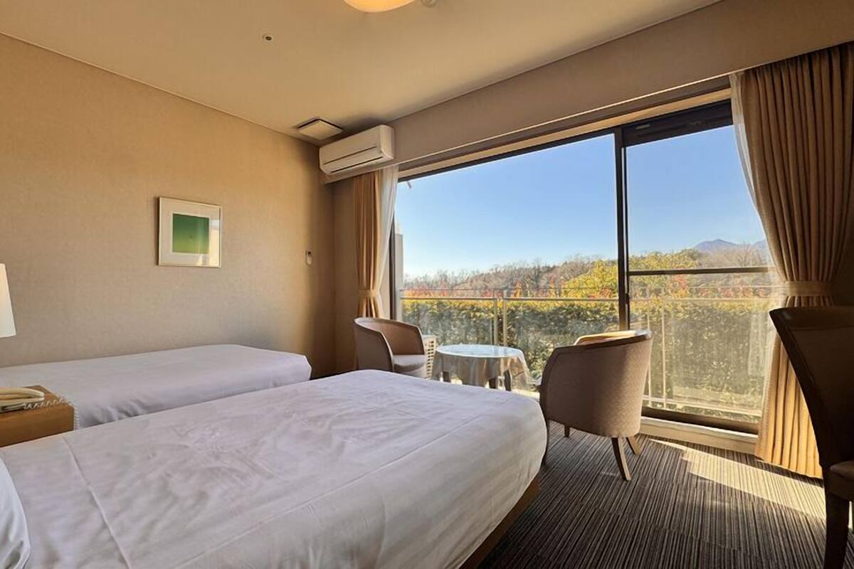 A hotel room with two beds, a desk, a chair, a small table, and a large window offering a view of trees and mountains.
