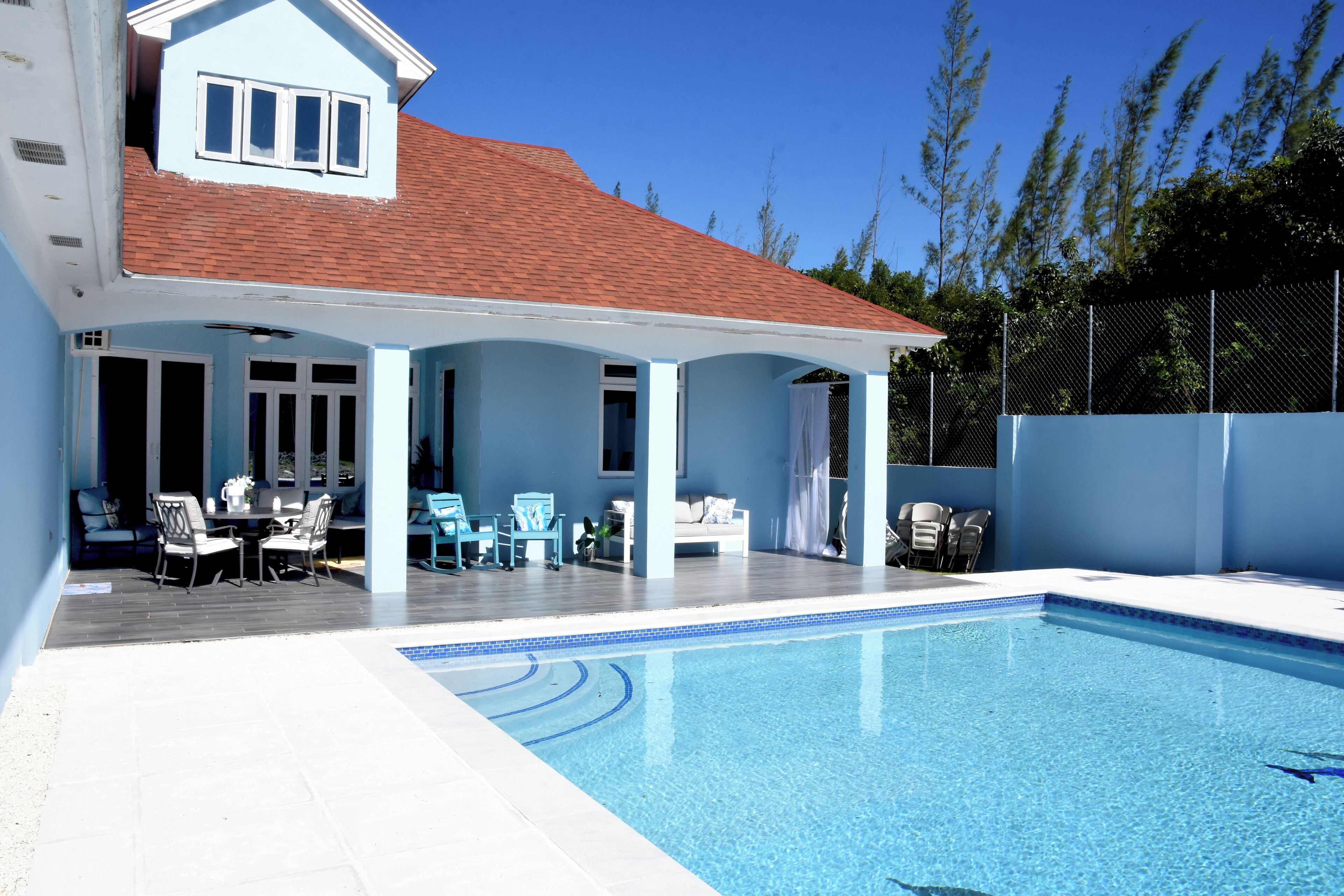 Ein Schwimmbad mit überdachter Terrasse, Sitzgelegenheiten im Freien und einem umzäunten Garten.