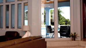 Un salon moderne avec un canapé, une lampe et une vue sur une piscine et un espace de détente extérieur, le tout visible à travers des portes vitrées.