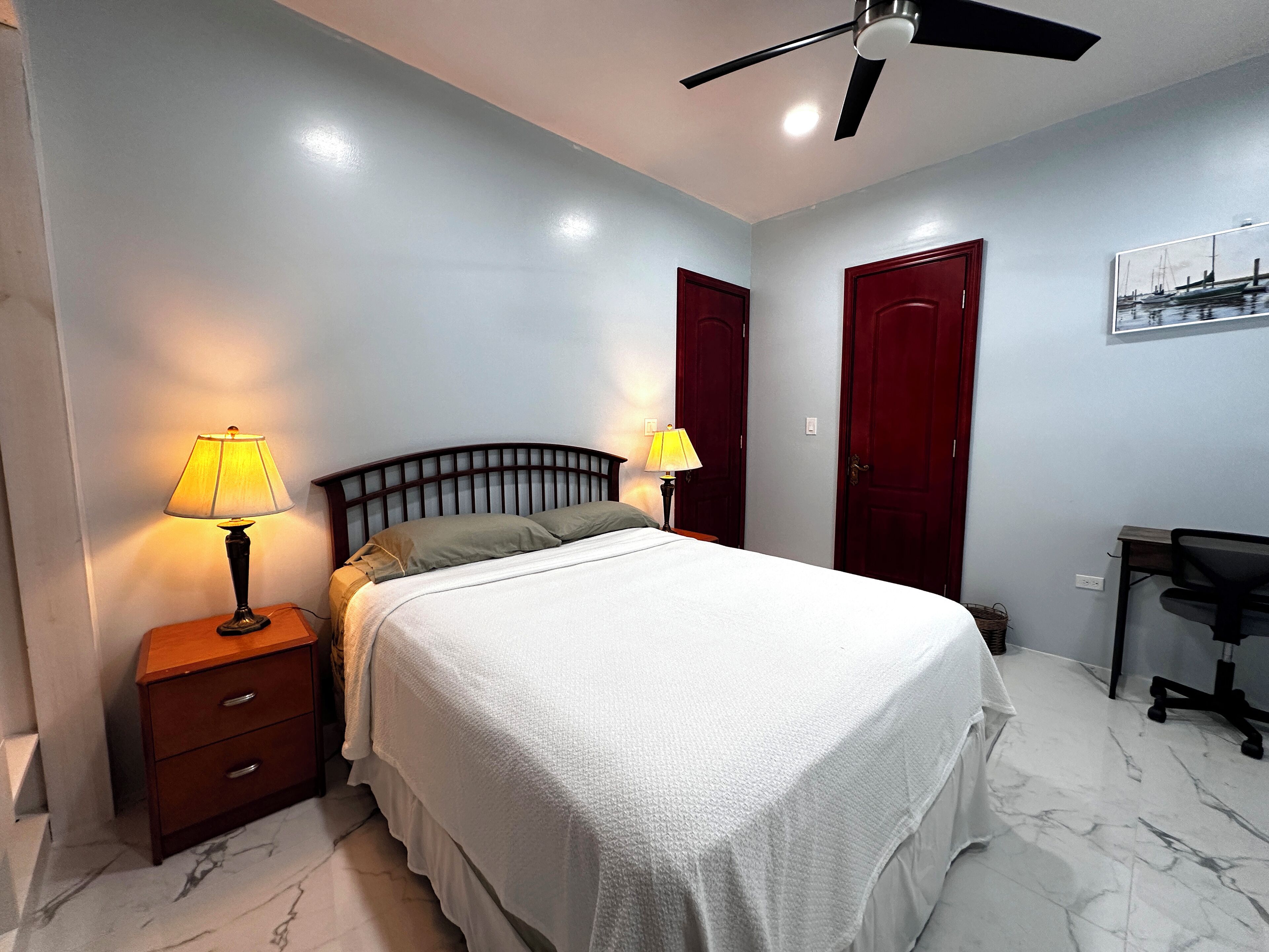 A neatly arranged bedroom with a bed, two bedside lamps, a desk, and a ceiling fan.