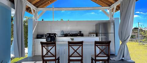 A covered outdoor kitchen with a wooden ceiling, white tiled backsplash, and a stainless steel refrigerator. There are three wooden chairs with white cushions and a small table.