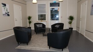 A lobby with black leather chairs, a small round table, and a white rug. There are two potted plants and a wall-mounted light fixture. - Britz Hostel (Kristinehamn)