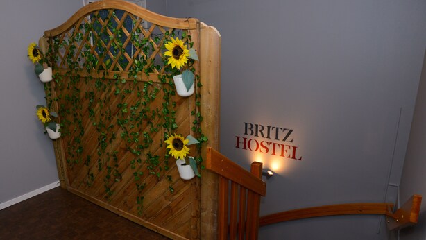 A wooden gate with a lattice design and sunflowers, leading to a staircase with a wooden handrail. The wall behind the gate has a sign that reads "BRITZ HOSTEL". - Britz Hostel (Kristinehamn)