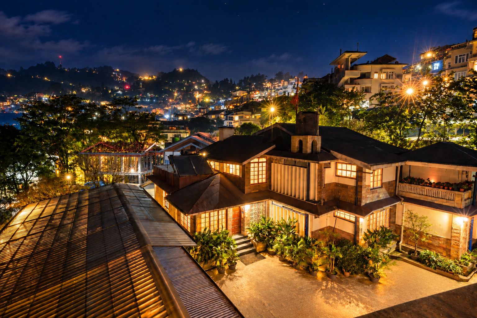 A multi-story house with a balcony, surrounded by potted plants, and a view of a cityscape at night.