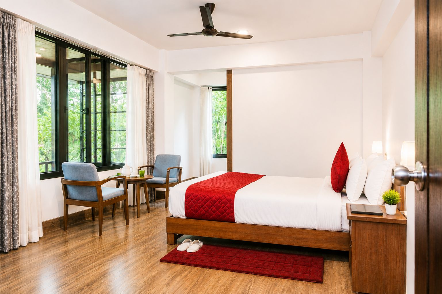 A hotel room with a bed, a desk, a chair, and a view of greenery through the windows.