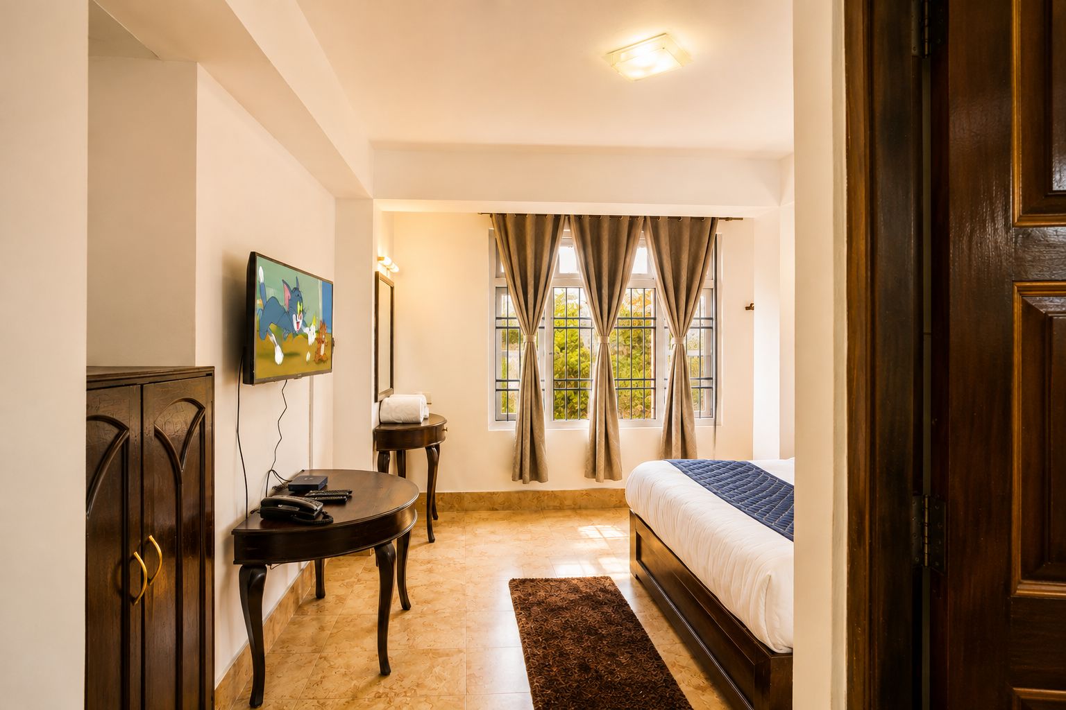 A hotel room with a bed, a television mounted on the wall, a small table, and a window with curtains.