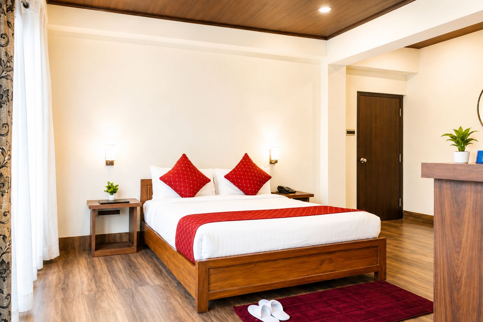 A hotel room with a wooden bed, red pillows, a red throw blanket, a nightstand with a lamp, a small plant, and a red rug.