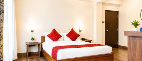 A hotel room with a wooden bed, red pillows, a red throw blanket, a nightstand with a lamp, a small plant, and a red rug.
