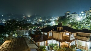 Una casa de varios pisos con balcón, rodeada de vegetación y vistas a un paisaje urbano nocturno.